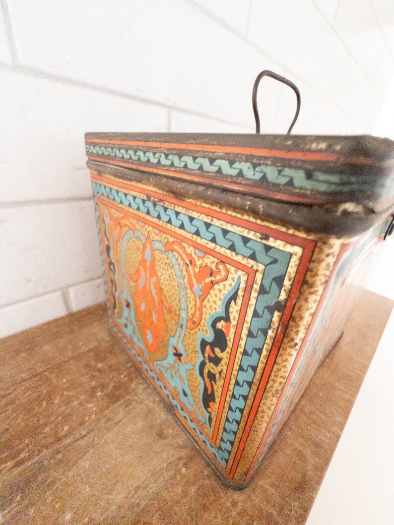 K&ouml;nnte beinhalten: Eine vintage Metalldose mit einem dekorativen Design mit komplizierten Mustern in Orange-, Blau- und Goldt&ouml;nen. Die Dose hat einen Klappdeckel und einen Drahtgriff.