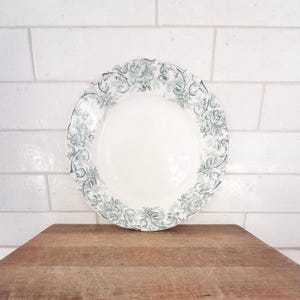 May include: A white ceramic plate with a detailed teal floral pattern around the rim. The plate is round with a slightly scalloped edge. The plate is set on a wooden surface against a white brick background.