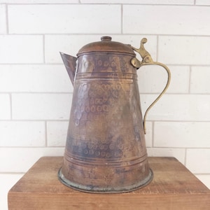 May include: A tall, tapered copper coffee pot featuring a hammered texture on the body, a dark patina, and a brass handle with a decorative thumb rest. The pot includes a matching lid with a small knob and a pointed spout, showing signs of age.
