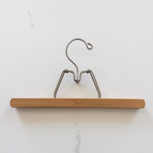 May include: A wooden clothes hanger with a metal hook and support arms. The wooden bar is light brown and has a small logo etched into it. The metal components are silver-colored.