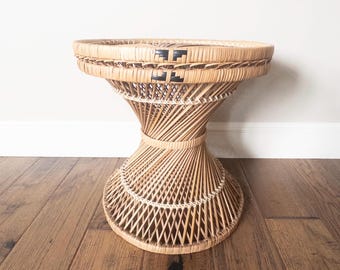 May include: A natural woven rattan side table with a unique hourglass shape. The table features a round top and a flared base, with black geometric accents. The table is approximately 20 inches in diameter and 20 inches tall.