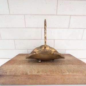 May include: A vintage brass basket with a decorative handle and ornate edges. The basket sits on a wooden cutting board against a white brick background. The basket is likely used for serving or display.