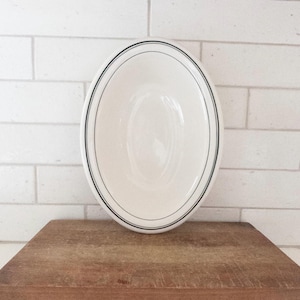 May include: An oval, off-white ceramic serving bowl with a thin black border. The bowl sits on a wooden cutting board against a white brick wall. The bowl's interior is smooth and reflective.