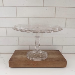 May include: A clear glass cake stand with a scalloped edge. The stand has a round base and a slender stem. The cake stand is sitting on a wooden surface.