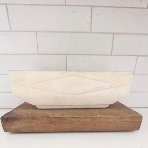 May include: A light-colored wooden bowl with a diamond pattern, resting on a rectangular wooden cutting board. The bowl has a rectangular shape with slightly angled sides. The cutting board is a dark brown color.