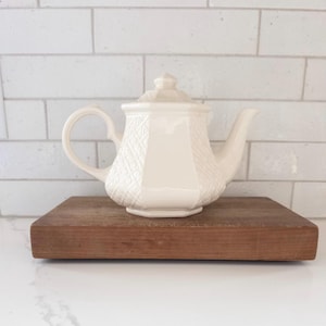 May include: A white ceramic teapot with a textured design sits on a wooden cutting board. The teapot has a rounded body and a spout on the side.