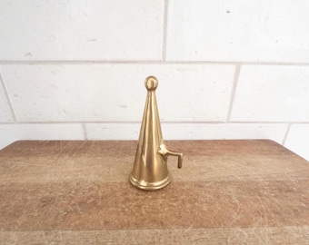 May include: A vintage brass funnel with a conical shape and a small spout. The funnel has a rounded top and is set on a wooden surface against a white brick wall. The brass has a polished, reflective finish.