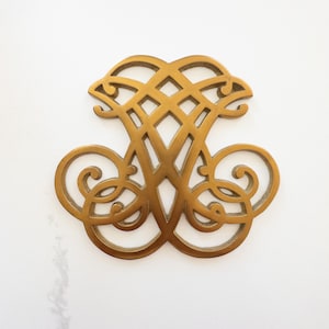 May include: A decorative, gold-colored metal object with an intricate, symmetrical design. The object features a central diamond-patterned section, flanked by swirling, scroll-like elements. The overall shape is reminiscent of a stylized letter or emblem.
