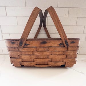 May include: A rectangular, woven brown basket with two arched wooden handles. The basket is made of interwoven strips of wood, with visible nail heads securing the handle. The basket is designed for carrying items.