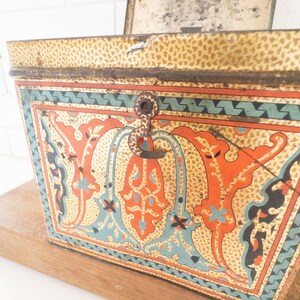 K&ouml;nnte beinhalten: Eine vintage Metalldose mit einem Scharnierdeckel und einem Verschluss. Die Dose ist mit einem farbenfrohen Blumenmuster in Orange-, Blau- und Gr&uuml;nt&ouml;nen verziert. Die Dose hat ein abgenutztes, gealtertes Aussehen.
