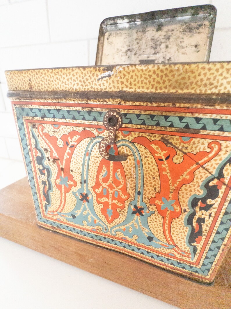 K&ouml;nnte beinhalten: Eine vintage Metalldose mit einem Scharnierdeckel und einem Verschluss. Die Dose ist mit einem farbenfrohen Blumenmuster in Orange-, Blau- und Gr&uuml;nt&ouml;nen verziert. Die Dose hat ein abgenutztes, gealtertes Aussehen.