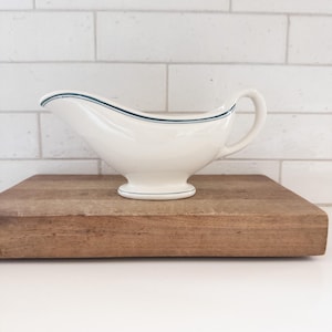 May include: A white ceramic gravy boat with a teal blue stripe detail. The gravy boat has a handle and a spout, and it sits on a wooden cutting board. The background is a white brick wall.