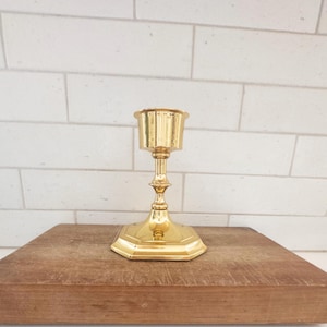 Könnte beinhalten: Ein goldfarbener Kerzenhalter mit sechseckigem Sockel und geriffelter Tasse. Der Kerzenhalter ist aus Metall gefertigt und hat ein klassisches Design. Er steht auf einer Holzoberfläche vor einer weißen Ziegelmauer.