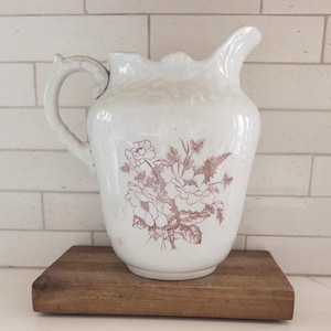 May include: White ceramic pitcher with a brown floral design. The pitcher has a handle and a spout. It is sitting on a wooden base.