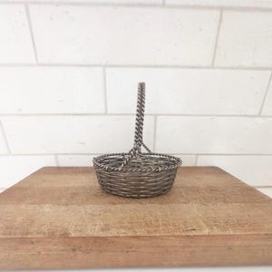 May include: A small, silver-toned wire basket with a woven design and a twisted handle. The basket is sitting on a wooden cutting board against a white brick wall. The basket is empty.