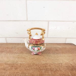 May include: A miniature porcelain teapot with a gold handle and accents. The teapot features a red and gold lid, a white spout, and a floral design on the body. The teapot is a decorative item.