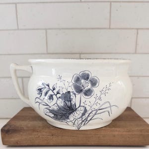 May include: A white ceramic bowl with a handle, decorated with a blue floral pattern. The bowl sits on a small, square wooden board. The floral design includes a large flower and leaves.