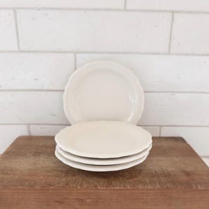 May include: A stack of four off-white ceramic plates with scalloped edges. The plates are round with a smooth, solid-colored surface and a slightly raised rim, featuring a consistent design across the entire set of dinnerware.