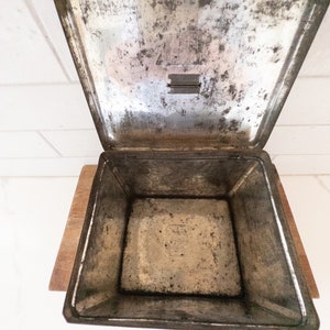 K&ouml;nnte beinhalten: Eine leere, silberne Metalldose mit einem Scharnierdeckel. Die Dose ist leicht angelaufen und hat eine kleine, rechteckige &Ouml;ffnung im Deckel.
