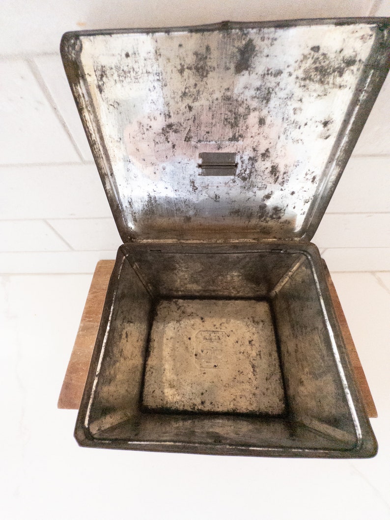 K&ouml;nnte beinhalten: Eine leere, silberne Metalldose mit einem Scharnierdeckel. Die Dose ist leicht angelaufen und hat eine kleine, rechteckige &Ouml;ffnung im Deckel.