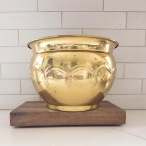 May include: A polished brass planter with a rounded, bulbous body and a wide, flared rim. The exterior surface features a repeating embossed scalloped pattern around the upper section, with a smooth, reflective finish throughout the metal.