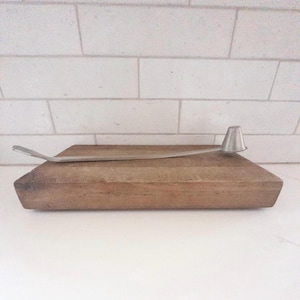 May include: A wooden cutting board with a stainless steel candle snuffer. The snuffer has a long, curved handle and a conical bell. The board is rectangular and made of light brown wood.