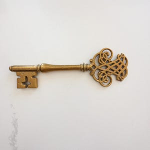 May include: A vintage-style brass key with an ornate, scrolled design on the head. The key has a rectangular bit and a detailed handle. The key is a warm gold color and lies on a white surface.