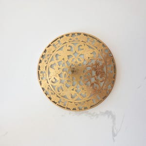 May include: A round, gold-colored decorative ceiling light fixture. The fixture features an intricate floral and vine pattern cut into the metal. The light shines through the openwork design, creating a decorative effect. The fixture is mounted on a white ceiling.