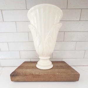 May include: Cream-colored ceramic vase with a fluted design and a pedestal base. The vase has a tulip-like shape and sits on a rectangular wooden cutting board. The background is a white brick wall.