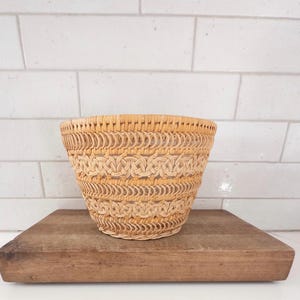 May include: A woven basket with a light brown color and a decorative pattern. The basket is sitting on a wooden cutting board. The background is a white tiled wall. This basket is ideal for home decor.