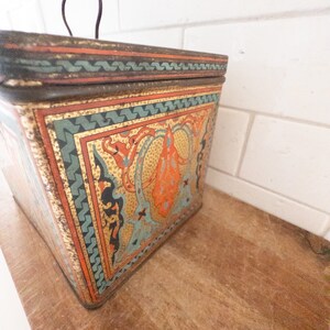 K&ouml;nnte beinhalten: Eine Vintage-Blechdose mit einem dekorativen Design in Gold, Orange, Blau und Gr&uuml;n. Die Dose hat einen Klappdeckel mit einem Metallgriff.