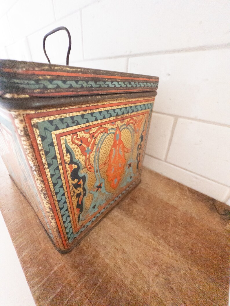 K&ouml;nnte beinhalten: Eine Vintage-Blechdose mit einem dekorativen Design in Gold, Orange, Blau und Gr&uuml;n. Die Dose hat einen Klappdeckel mit einem Metallgriff.
