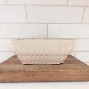 May include: A cream-colored, vintage-style bowl with a textured, ridged design. The bowl sits on a rectangular wooden cutting board. The background features white, rectangular tiles.