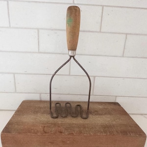 Könnte beinhalten: Ein Vintage-Kartoffelstampfer mit einem verwitterten Holzgriff, der einen kleinen grünen Farbklecks aufweist, und einem Kopf aus dunklem, oxidiertem Metalldraht in einer durchgehenden Wellenform. Das Werkzeug hat eine Metallzwinge.