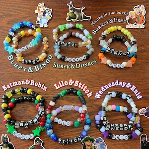 May include: A set of 10 colorful friendship bracelets with different cartoon character names, including Bluey & Bingo, Shrek & Donkey, Barney & Fred, Batman & Robin, Lilo & Stitch, and Wednesday & Enid. The bracelets are made with colorful plastic beads and star-shaped charms.