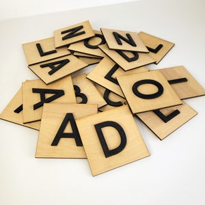 May include: A set of wooden letter tiles with black lettering. The tiles are arranged in a pile, with the letters A, B, C, D, E, F, G, H, I, J, K, L, M, N, O, P, Q, R, S, T, U, V, W, X, Y, and Z visible.