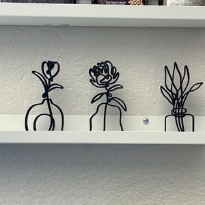 May include: Three black metal wire sculptures of flowers in vases on a white shelf. The flowers are stylized and abstract.