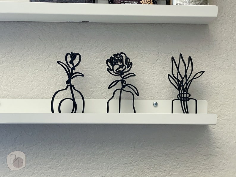 May include: Three black metal wire sculptures of flowers in vases on a white shelf. The flowers are stylized and abstract.