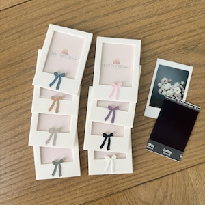 May include: White photo frames with colorful bows, including blue, tan, gray, and green, are stacked on a wooden surface. Each frame has a light pink interior and the text "PRINT PURPOSEFULLY". A photo and Instax film are also present.
