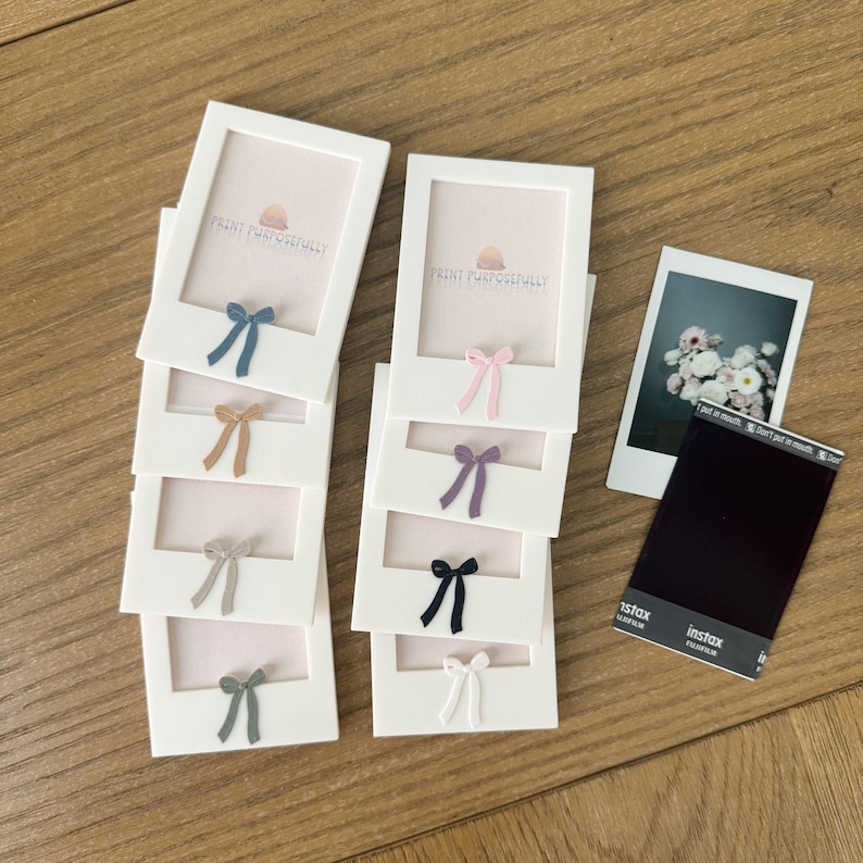 May include: White photo frames with colorful bows, including blue, tan, gray, and green, are stacked on a wooden surface. Each frame has a light pink interior and the text "PRINT PURPOSEFULLY". A photo and Instax film are also present.