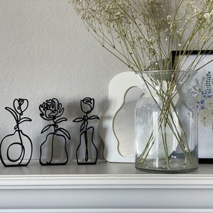 May include: Three black metal wire sculptures of flowers in vases on a white mantel. The flowers are stylized and abstract. The vases are simple and modern.