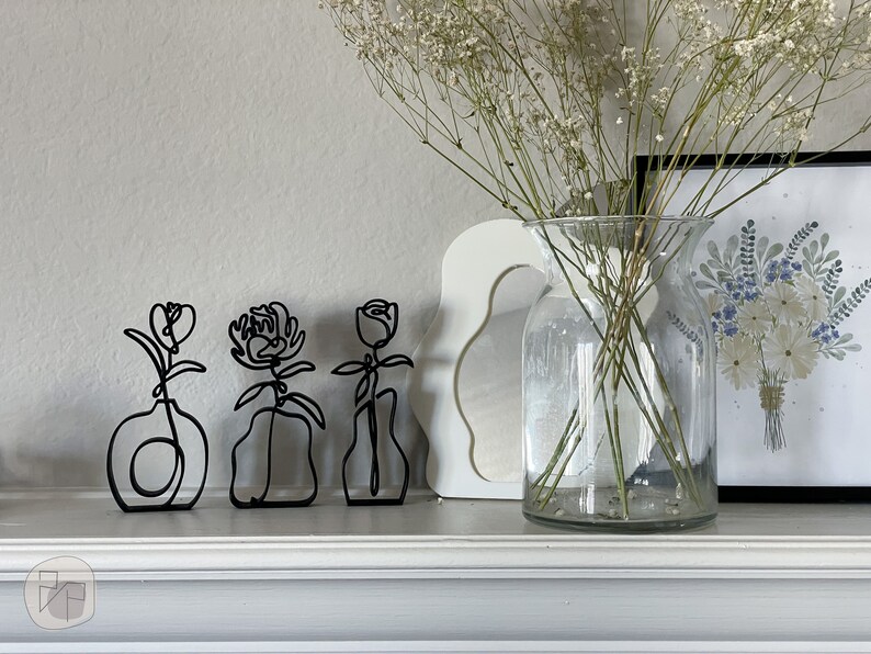 May include: Three black metal wire sculptures of flowers in vases on a white mantel. The flowers are stylized and abstract. The vases are simple and modern.
