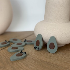 Puede incluir: Un conjunto de adornos verdes en forma de aguacate con huesos marrones y ojos negros. Los adornos están dispuestos sobre una superficie de madera junto a un jarrón de cerámica beige.