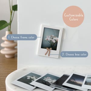 May include: A white photo frame with a small bow at the bottom, displaying a photo of flowers. The text "Customizable Colors" is in a peach circle. The text "1. Choose frame color" and "2. Choose bow color" are also visible.