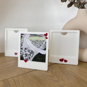 May include: Three white Polaroid-style photo frames, each with a small red heart detail. One frame displays a wedding veil photograph. The frames are on a wooden surface, with a vase and dried foliage in the background.