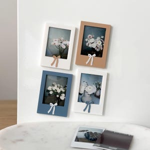 May include: Four rectangular photo frames in white, tan, and blue, each with a small bow at the bottom. The frames hold images of flowers in vases. A polaroid photo with a leaf design is in the foreground.