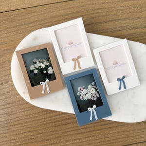 Puede incluir: Cuatro marcos de fotos rectangulares en tonos marrón, blanco y azul, cada uno con un pequeño lazo. Dos marcos contienen arreglos florales, mientras que los otros dos tienen el texto "PRINT PURPOSEFULLY". Los marcos están dispuestos sobre una superficie blanca de forma ovalada.
