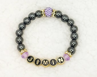 Könnte beinhalten: Ein Armband mit dunkelgrauen, runden Perlen. Das Armband enthält goldfarbene Akzente, lila Kristallperlen und schwarze und goldene Buchstabenperlen mit der Aufschrift "JIMIN". Eine große, lila Strassperle ist mittig angeordnet.
