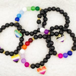 May include: A collection of five stretch bracelets featuring matte black beads interspersed with segments of colorful beads. Each bracelet includes a gold-toned metal charm shaped like a heart, displaying various pride flag color patterns.