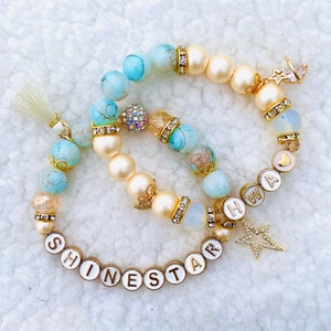 May include: Two bracelets with white beads, blue and white marbled beads, and gold charms. One bracelet has the word "SHINESTAR" spelled out in gold letters on white beads. The other bracelet has the word "HAWAII" spelled out in gold letters on white beads.